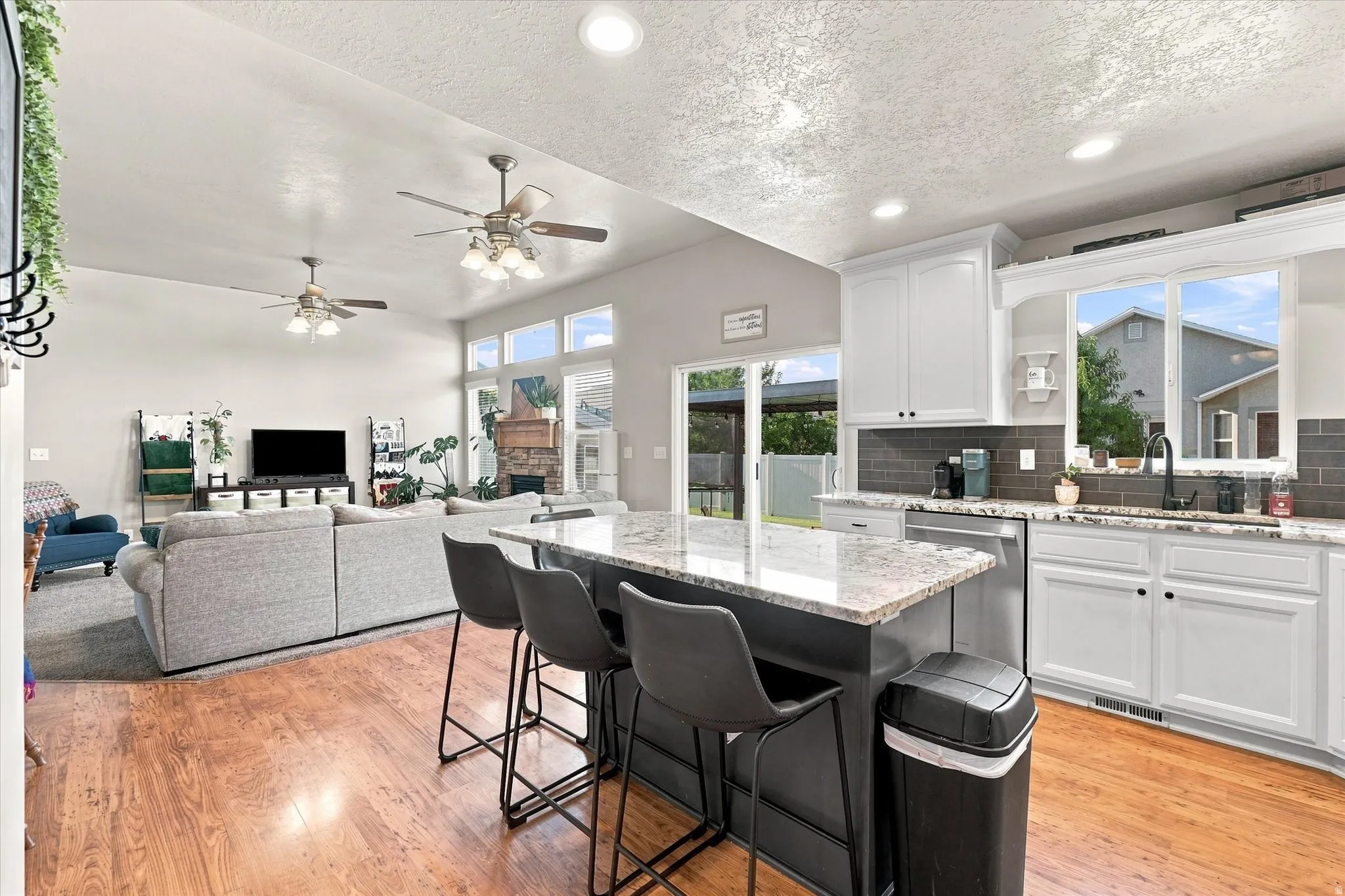 Open-concept living area with vaulted ceilings, wood-finish flooring, a central kitchen island with seating, white cabinetry, and a stone fireplace
