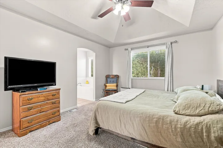 Vaulted ceiling with ceiling fan, carpet flooring, window with blinds, and an arched doorway leading to a built-in tub