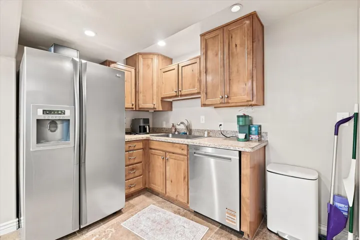 Stainless steel side-by-side refrigerator, stainless steel dishwasher, light wood-finish cabinetry, speckled countertop, and recessed lighting