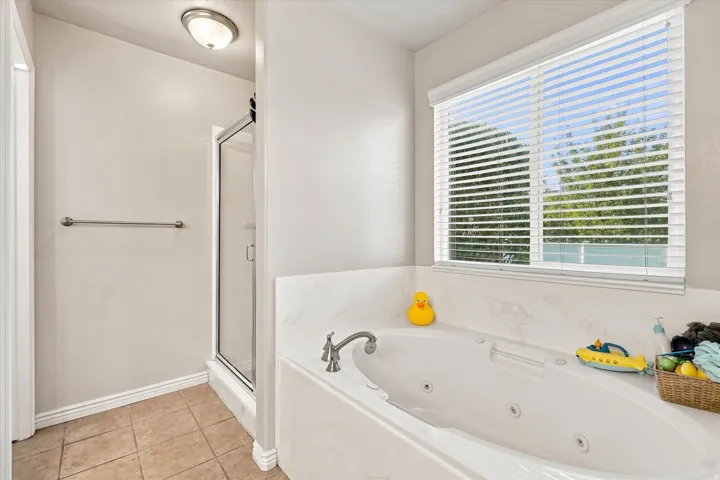 Soaking tub with integrated jets, tiled flooring, glass-enclosed shower, and a window with blinds
