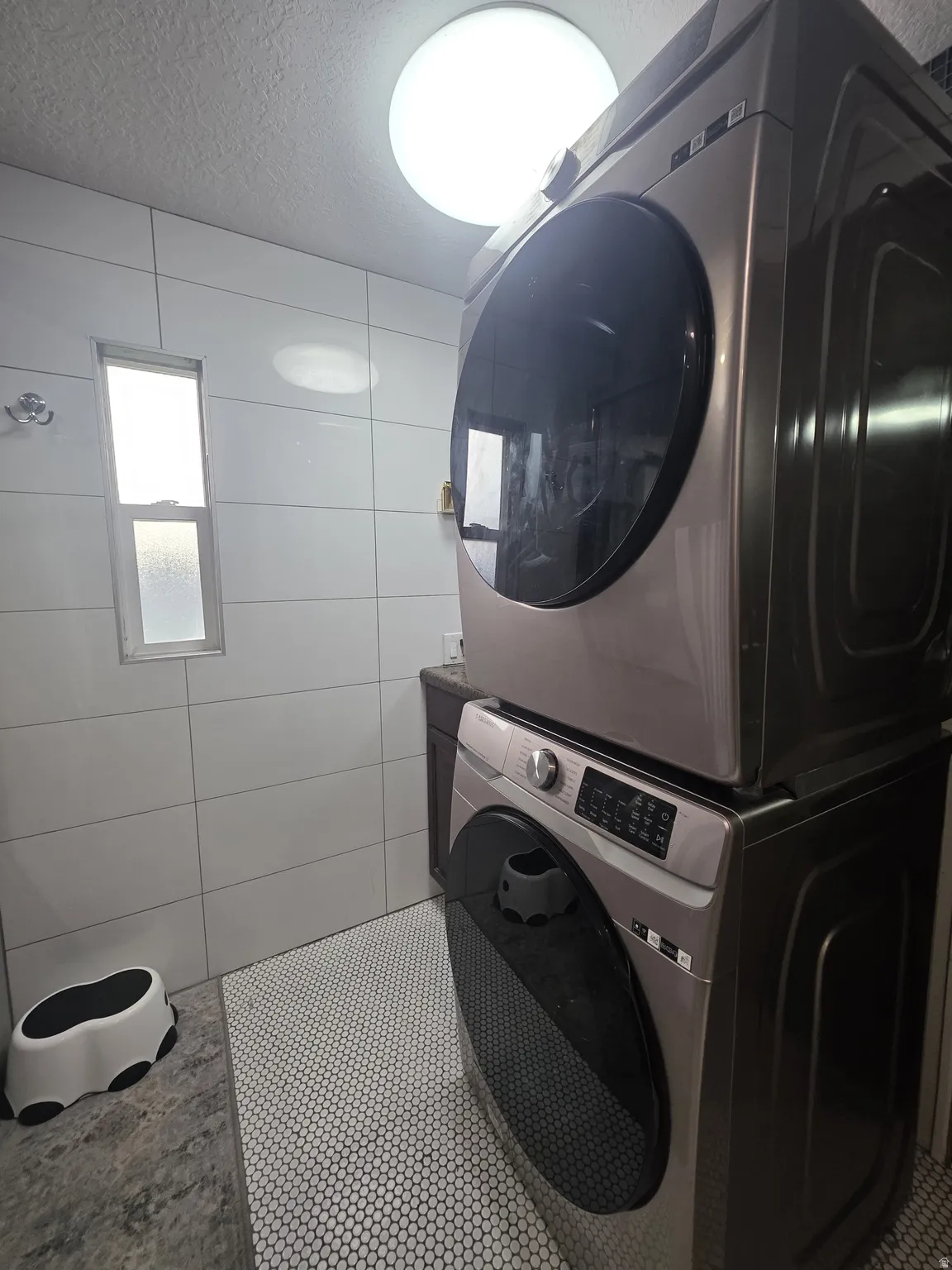 Stacked washer and dryer units, white tiled walls, white penny tile flooring, recessed window, and flush-mount ceiling light