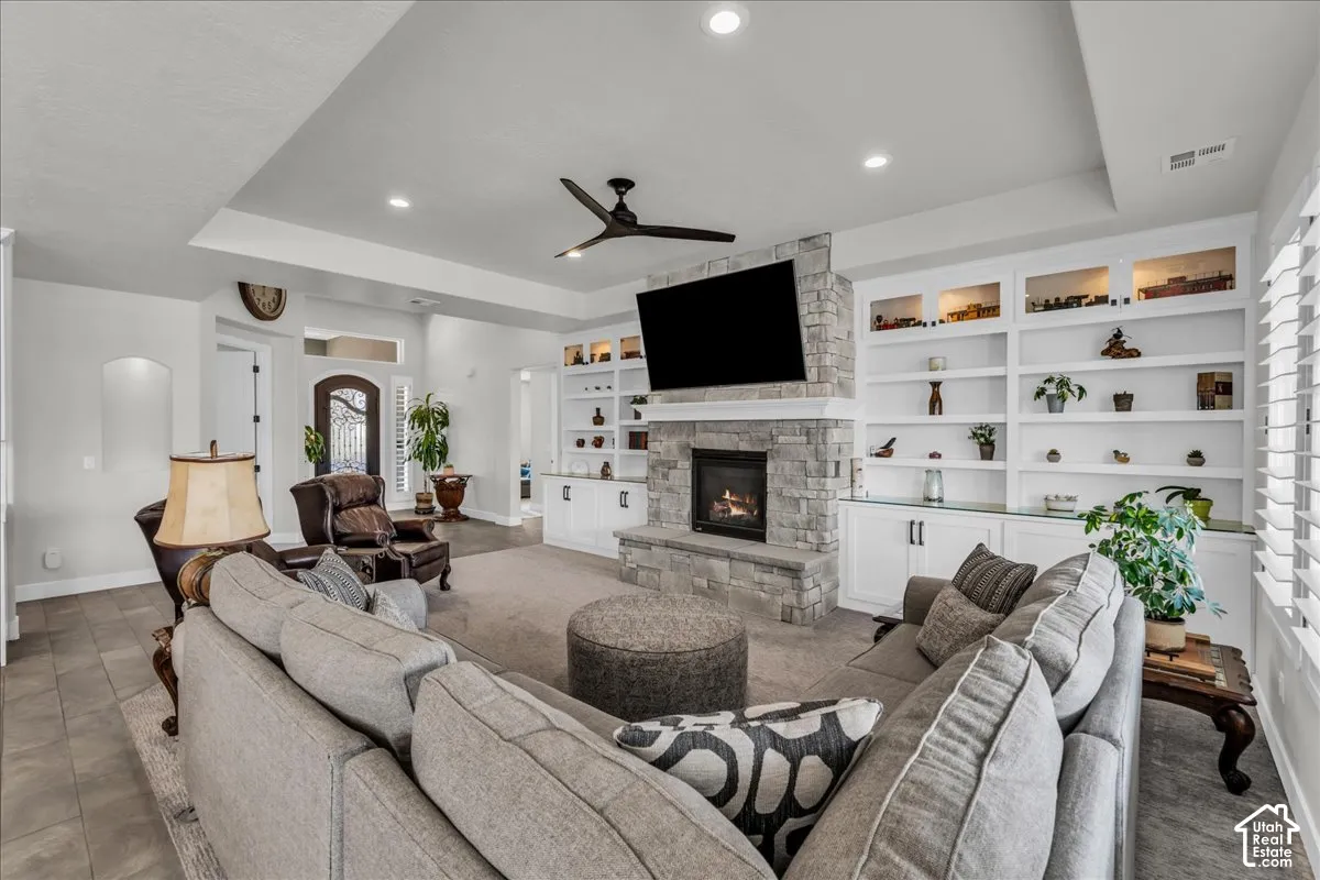 Living area featuring a stone fireplace, integrated built-in shelving, tray ceiling, recessed lighting, and a ceiling fan