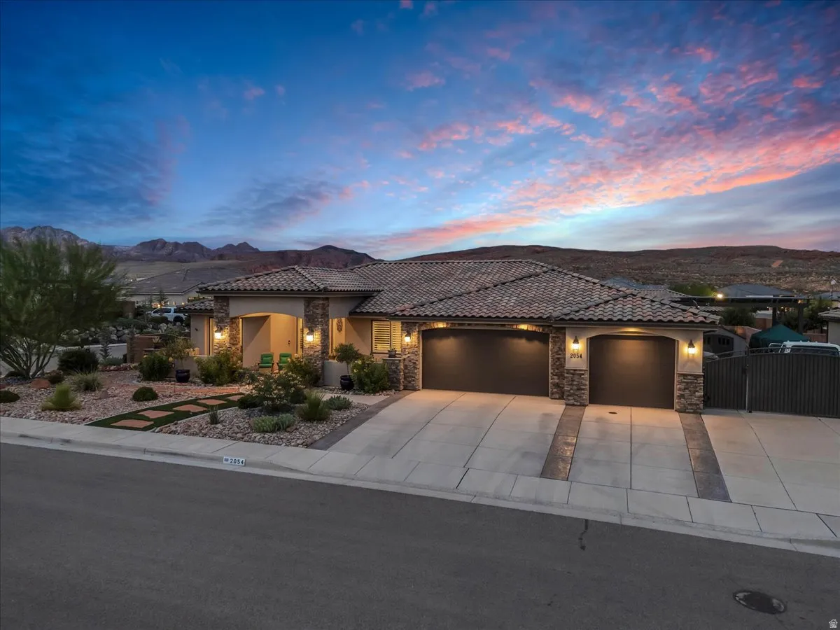 Stunning contemporary residence featuring a tile-and-stone exterior with architectural detail, dual three-car garage doors with dark finishes, professional landscape lighting throughout the front elevation, and manicured desert landscaping with mature veg