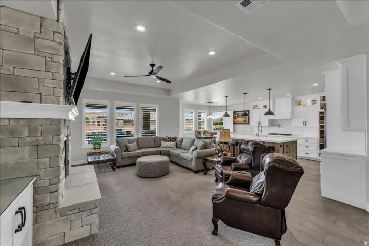 Stone-clad fireplace with mantel, recessed lighting, ceiling fan, white kitchen cabinetry, and pendant lighting