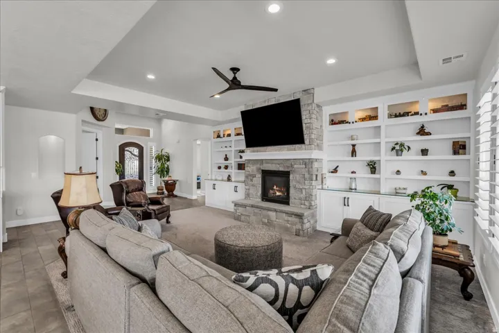 Living area featuring a stone fireplace, integrated built-in shelving, tray ceiling, recessed lighting, and a ceiling fan