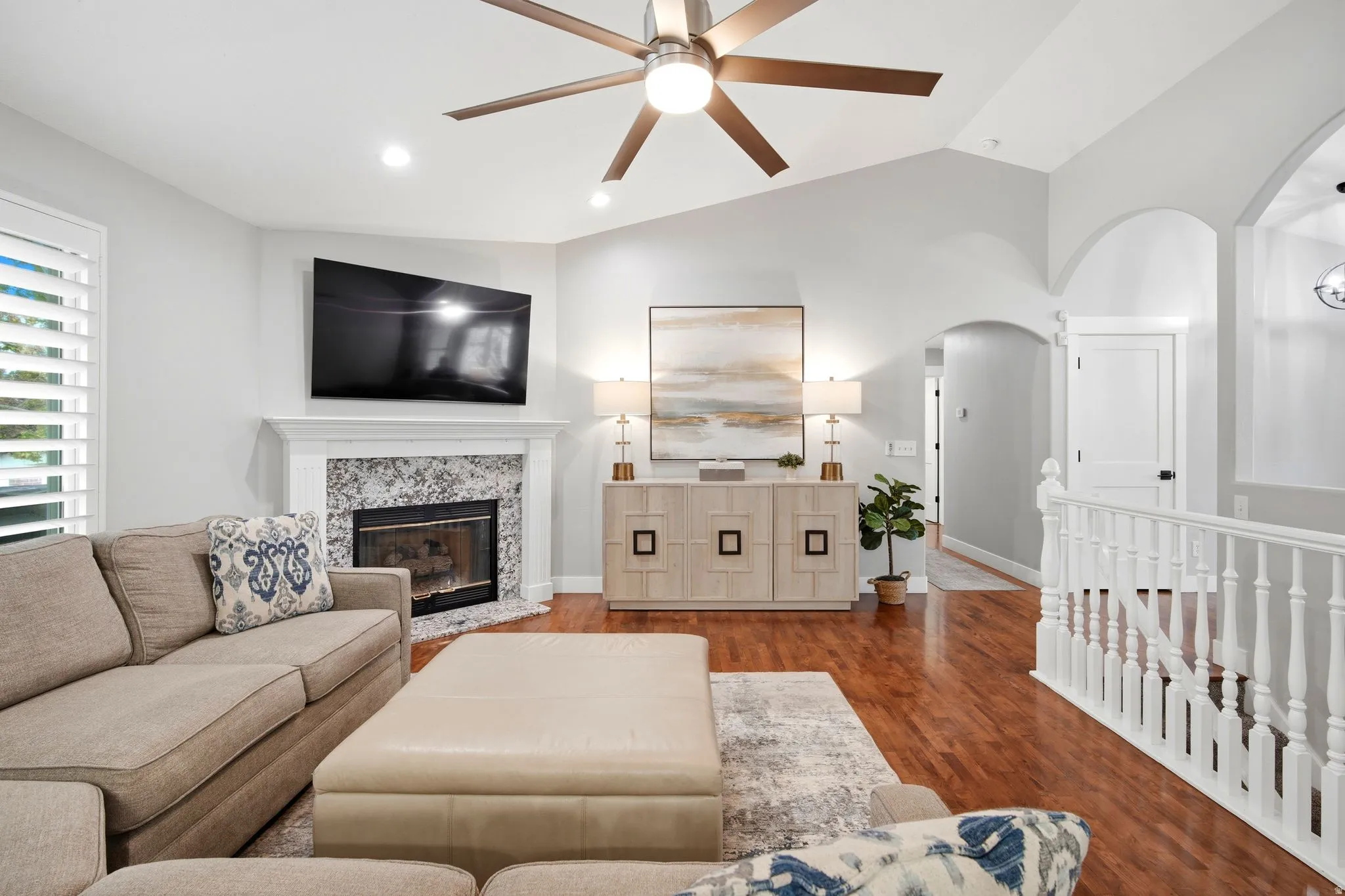 Bright open-concept living area featuring hardwood flooring, a gas fireplace with stone surround and built-in cabinetry, ceiling fan with integrated lighting, and large flat-screen television mount