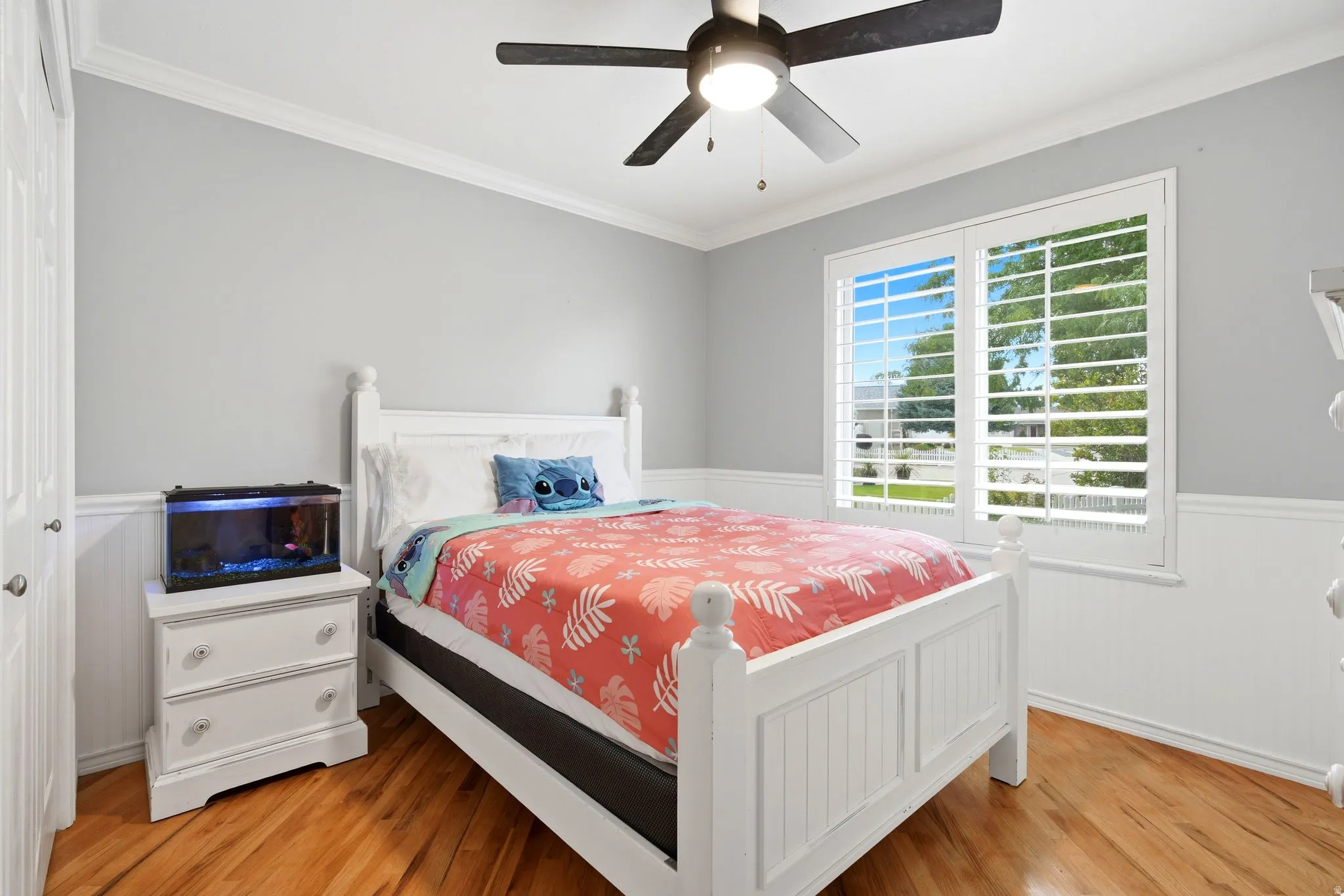 Bright secondary bedroom featuring hardwood flooring, white painted wood paneling, a ceiling fan with integrated lighting, and plantation shutter-style window treatments allowing natural light and privacy control