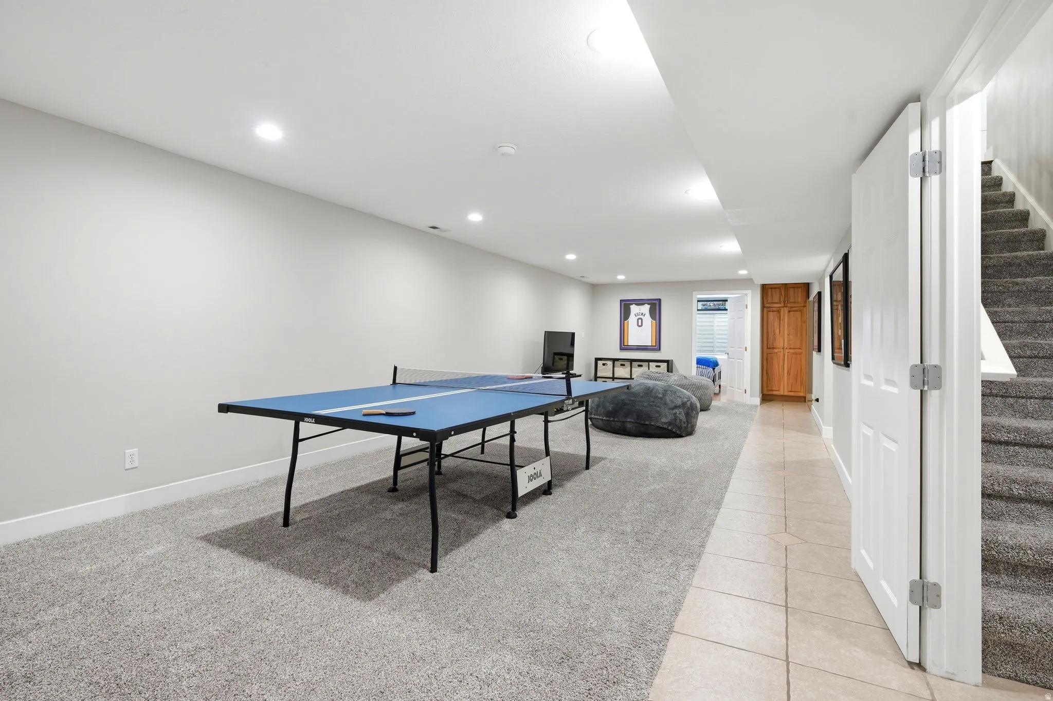 Finished basement recreation area featuring a blue regulation ping pong table, neutral tile flooring, recessed ceiling lighting, and white trim framing