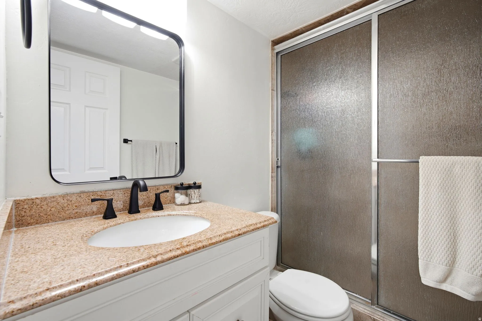 Spa-inspired bathroom featuring a framed mirror with integrated lighting, dual-handle faucet with matte black finish, and warm stone-finish countertop with an undermount sink