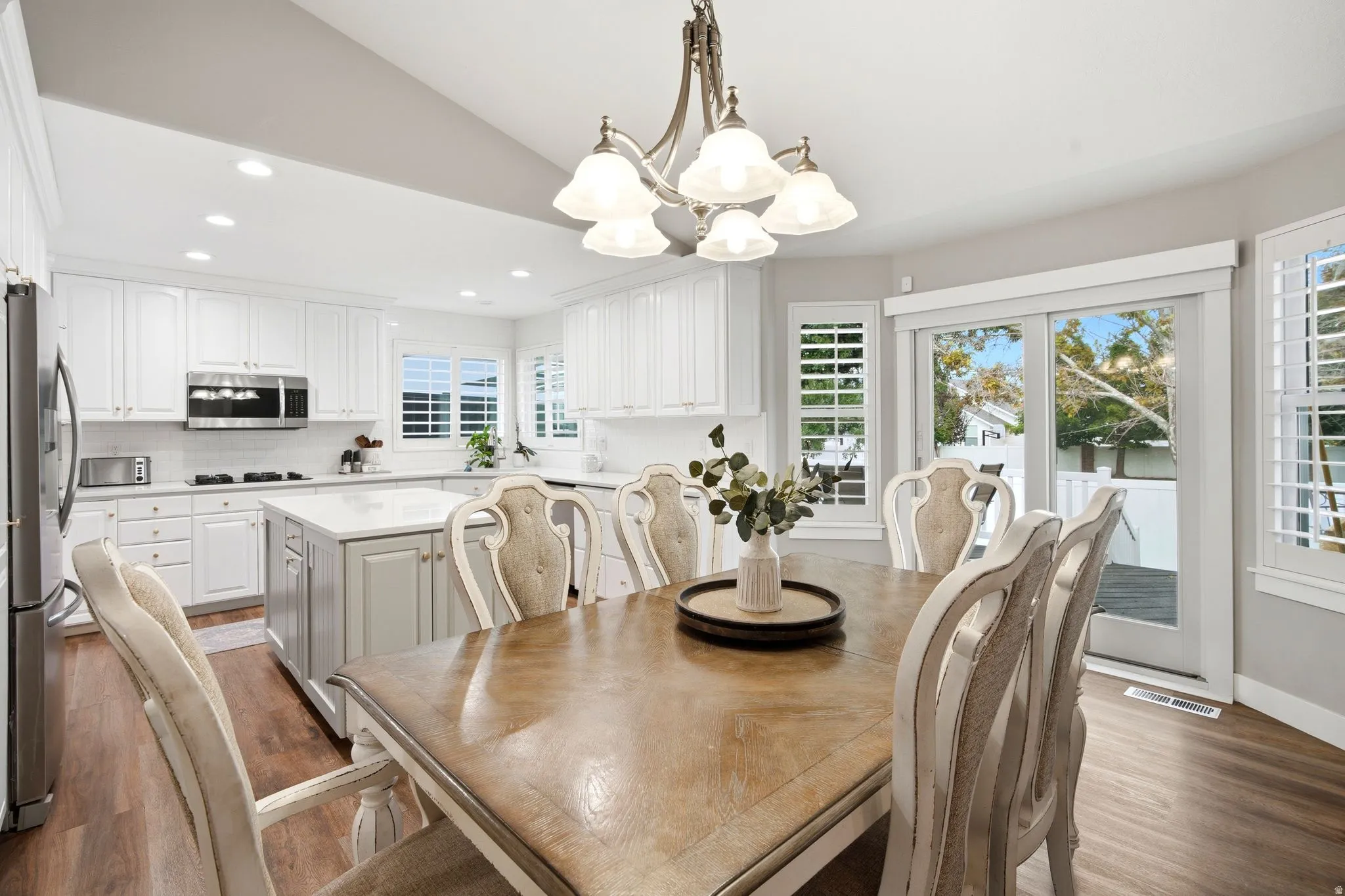 Open-concept kitchen and dining area featuring white cabinetry, stainless steel appliances, quartz countertops, a wood-finish dining table with seating for six, recessed ceiling lighting, and glass doors opening to outdoor space