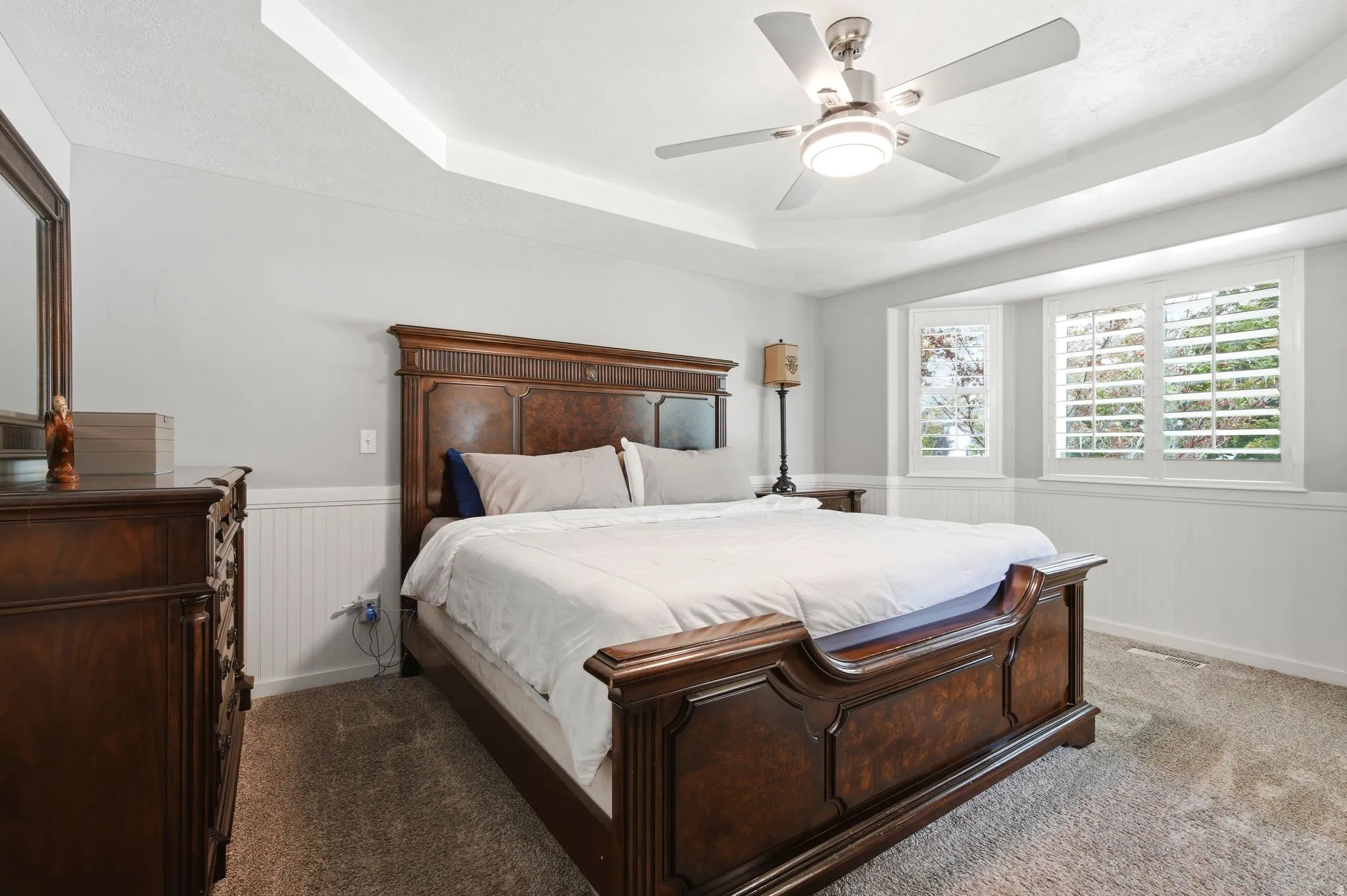 Tray ceiling with a ceiling fan, bay window with plantation shutters, and beadboard wainscoting