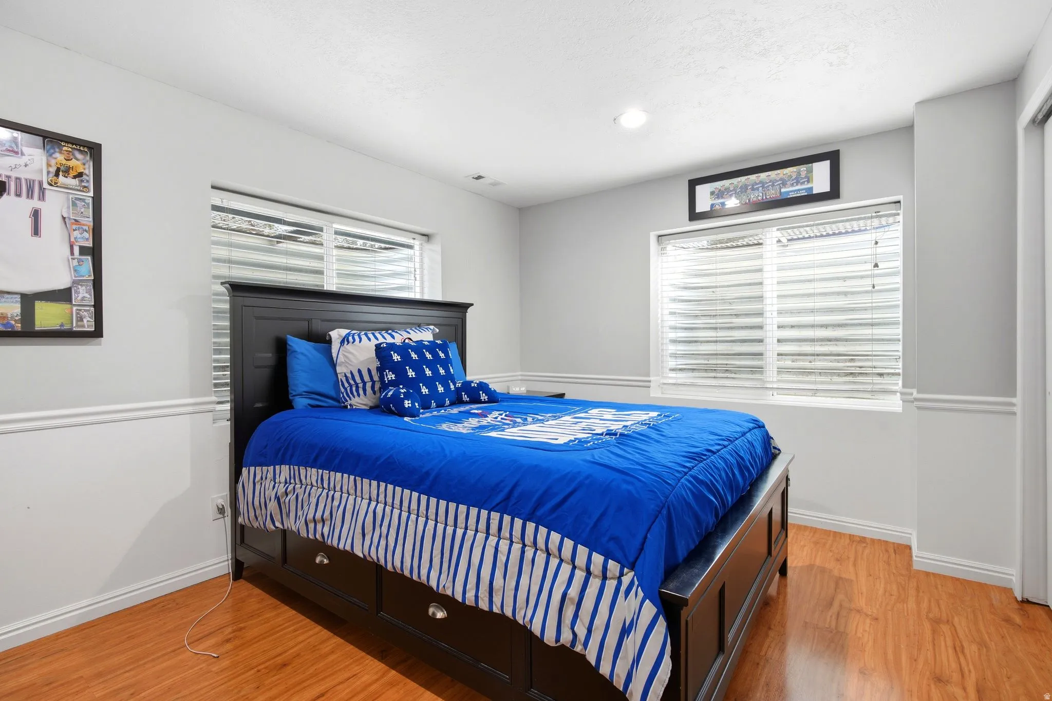 Bright secondary bedroom featuring warm wood-finish flooring, black metal bed frame, blue bedding ensemble, plantation shutters on dual windows with natural light, white wainscoting detail, and recessed ceiling lighting