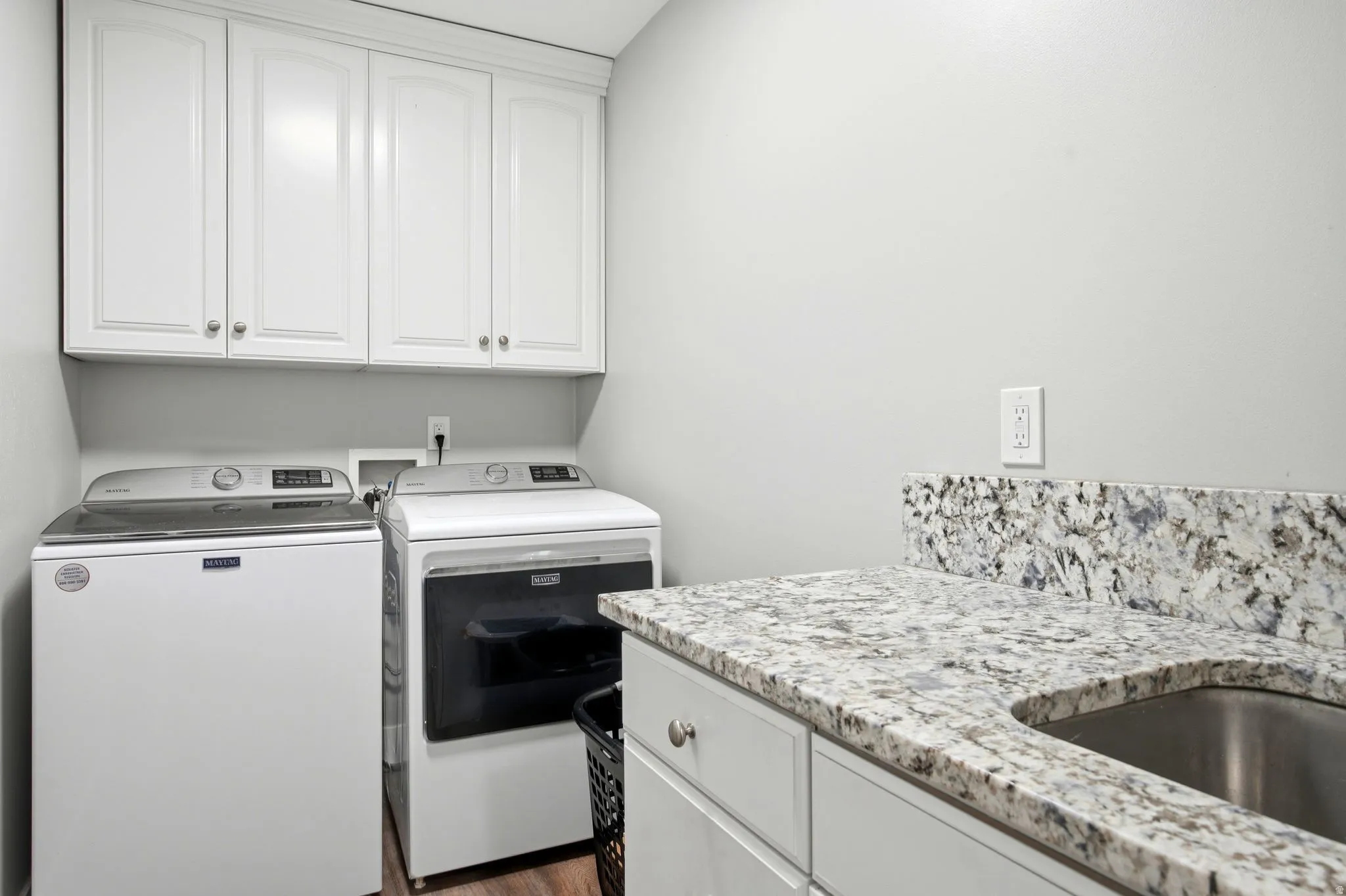 Modern laundry room featuring stacked front-load washer and electric dryer, white cabinetry with brushed nickel hardware, speckled granite-style countertop with undermount stainless steel sink, and abundant storage above