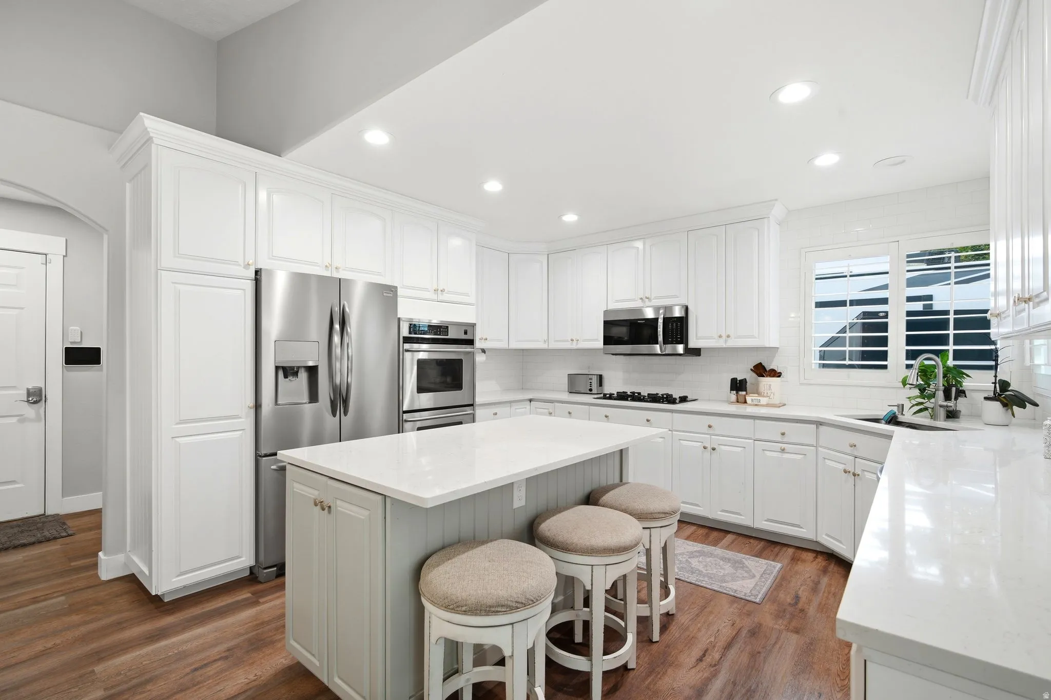 Chef's kitchen featuring white cabinetry with shaker-style doors, a large center island with seating, stainless steel appliances including a French-door refrigerator and dual wall ovens, quartz countertops, recessed ceiling lighting, and wood-finish floor