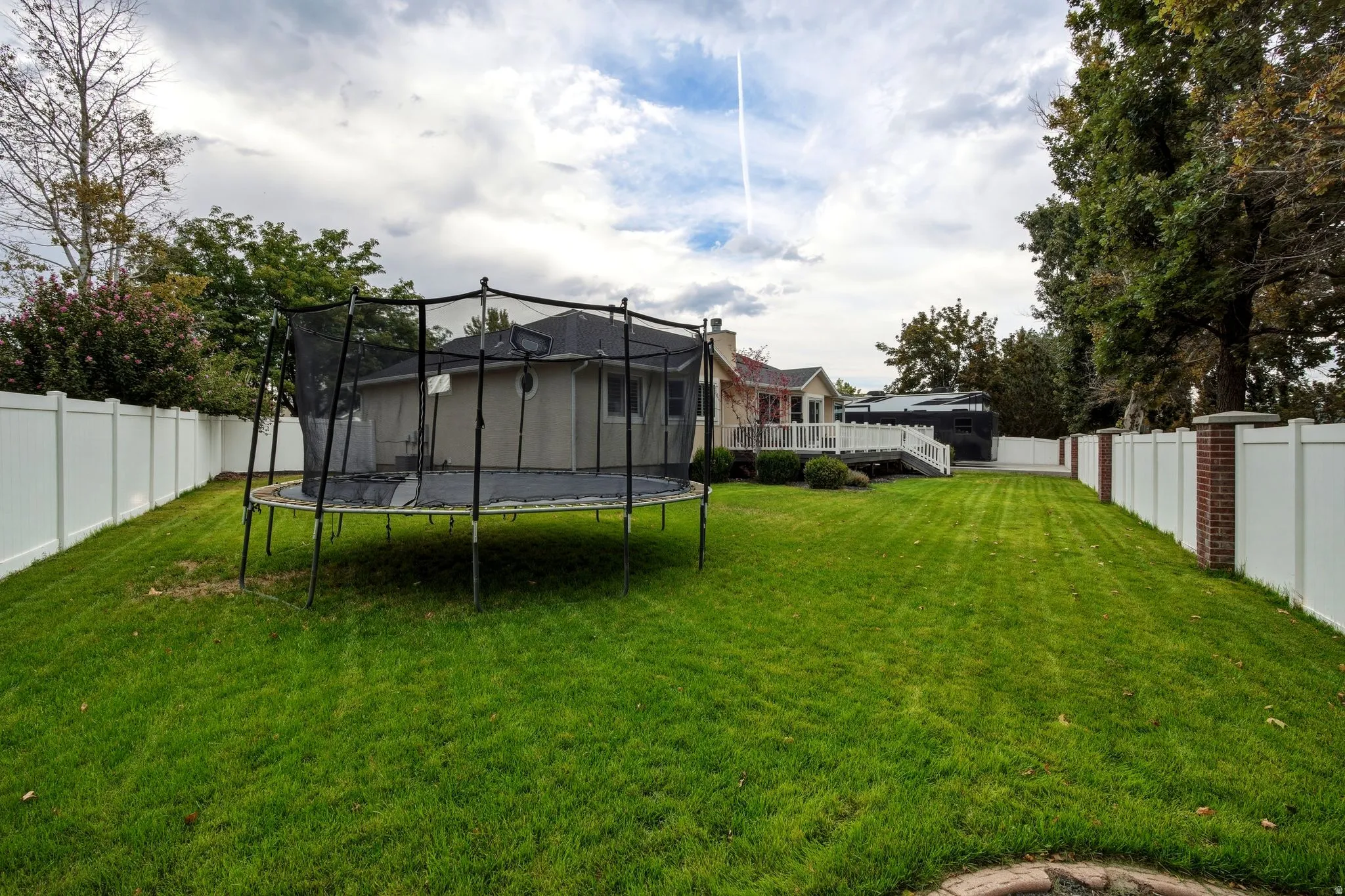 Expansive backyard featuring a meticulously manicured lawn, mature landscaping with established trees lining the perimeter, white vinyl fencing, and a black trampoline with safety enclosure