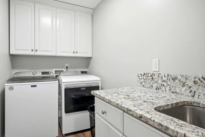 Modern laundry room featuring stacked front-load washer and electric dryer, white cabinetry with brushed nickel hardware, speckled granite-style countertop with undermount stainless steel sink, and abundant storage above