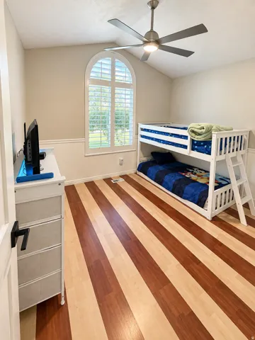Arched window with plantation shutters, vaulted ceiling, ceiling fan with integrated lighting, two-tone striped wood-finish flooring, and crown molding