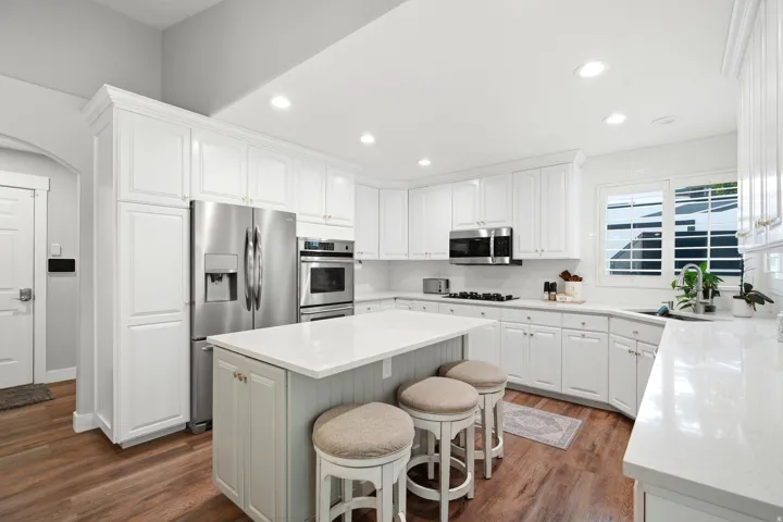 Chef's kitchen featuring white cabinetry with shaker-style doors, a large center island with seating, stainless steel appliances including a French-door refrigerator and dual wall ovens, quartz countertops, recessed ceiling lighting, and wood-finish floor
