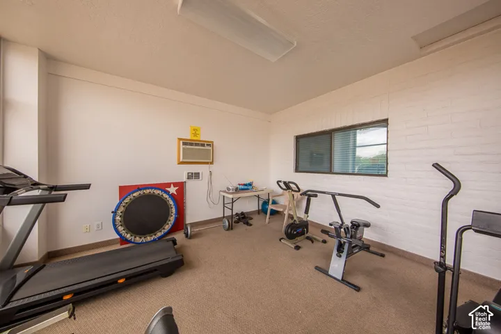 Carpeted floor, painted brick accent wall, white painted walls, window with blinds, and fluorescent ceiling lighting