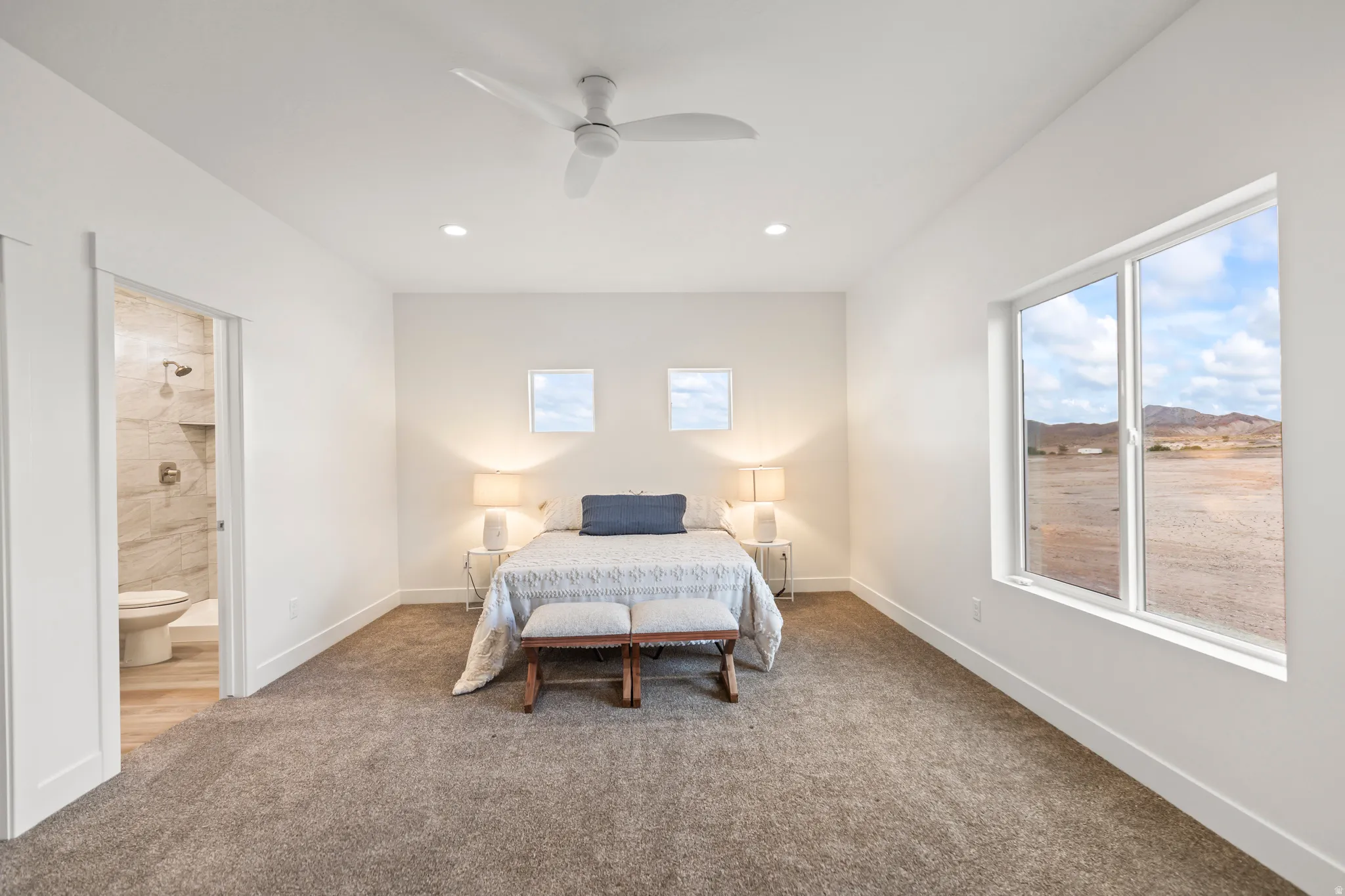 Recessed lighting, ceiling fan, expansive window, plush carpet flooring, and an adjoining bathroom with a tile-surround shower