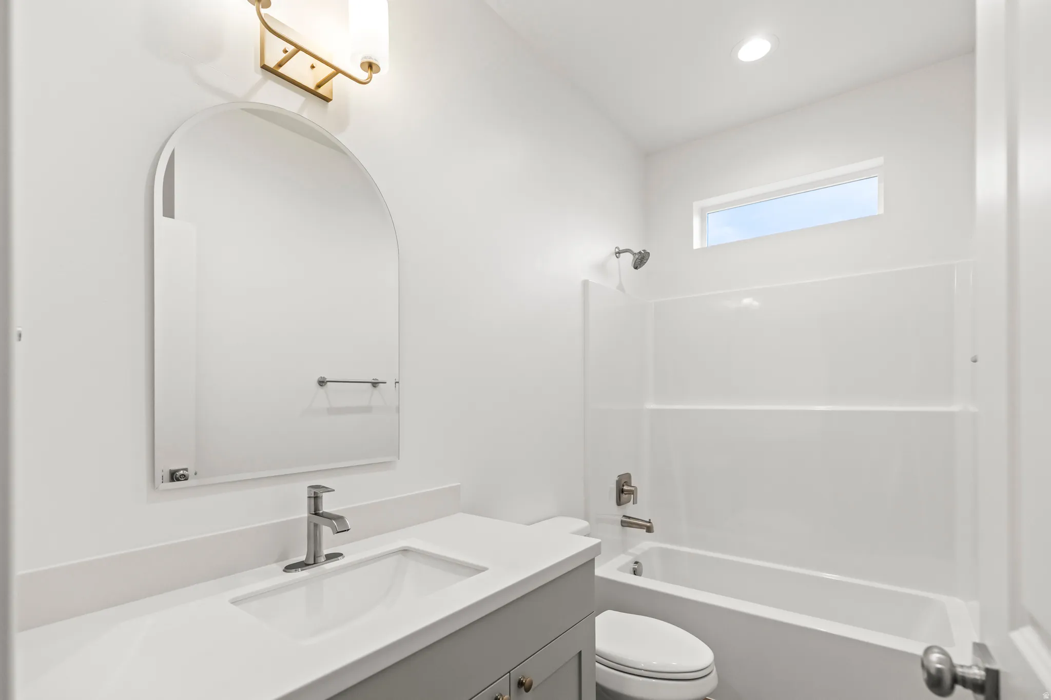 White single vanity with an integrated sink and brushed nickel faucet, arched mirror, recessed lighting, and a bathtub with a shower and window