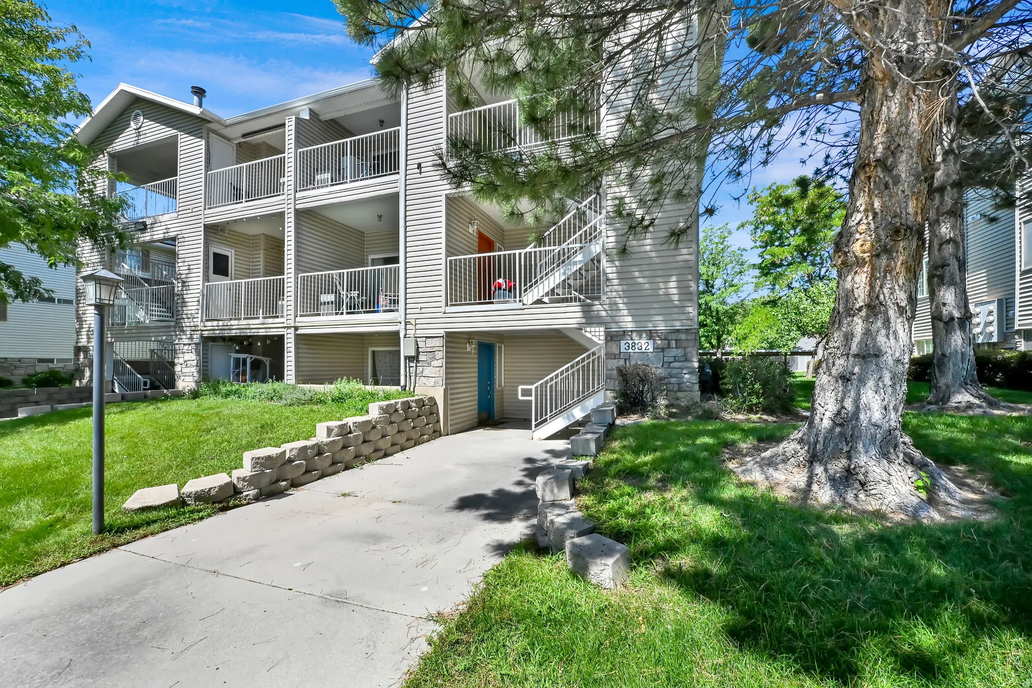 Multi-story building featuring stacked balconies with white railings, a paved drive, and stone retaining walls