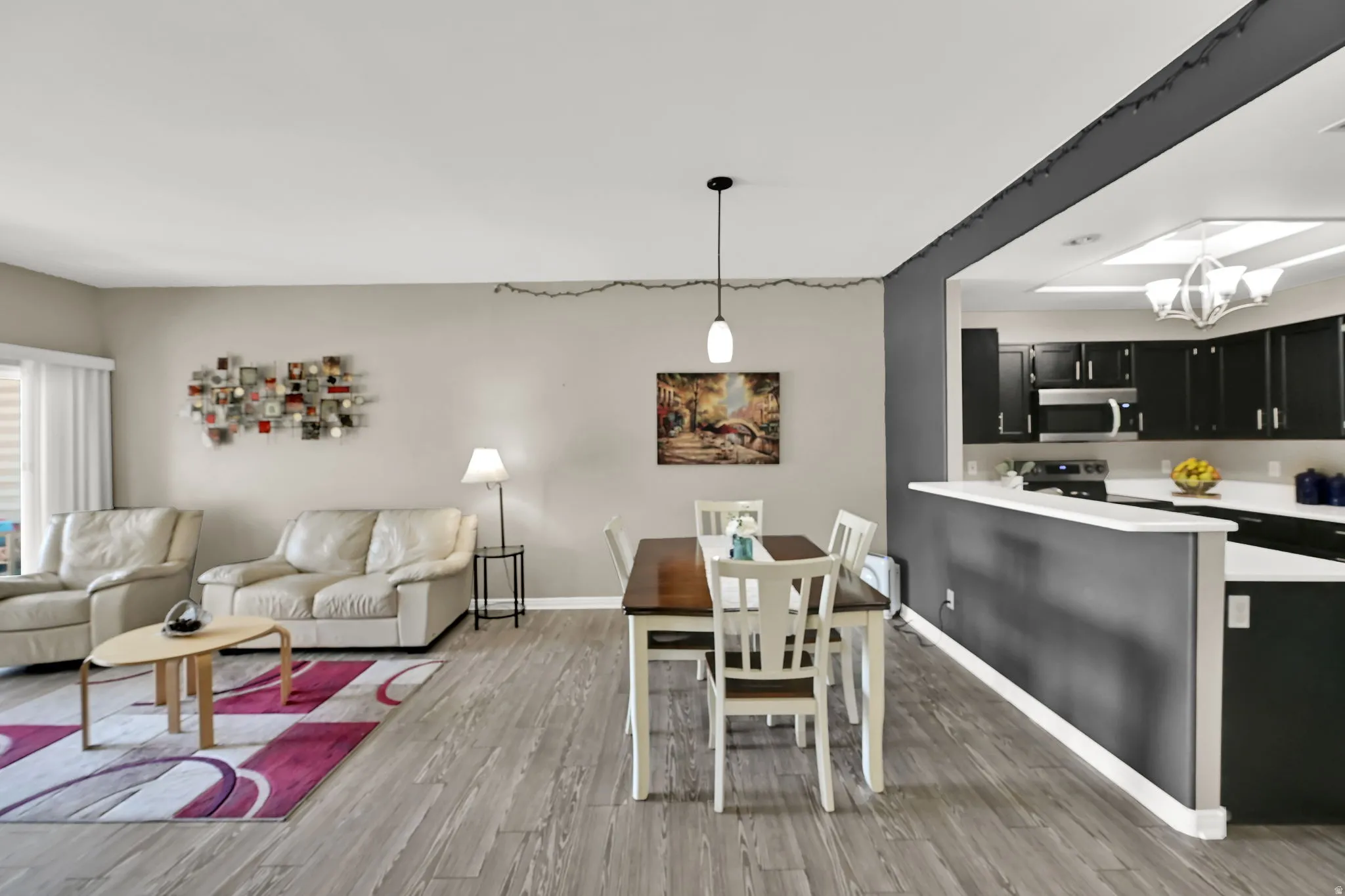 Open-concept living area with wood-finish flooring, a dining space featuring a pendant light fixture, and a kitchen with dark cabinetry and light countertops