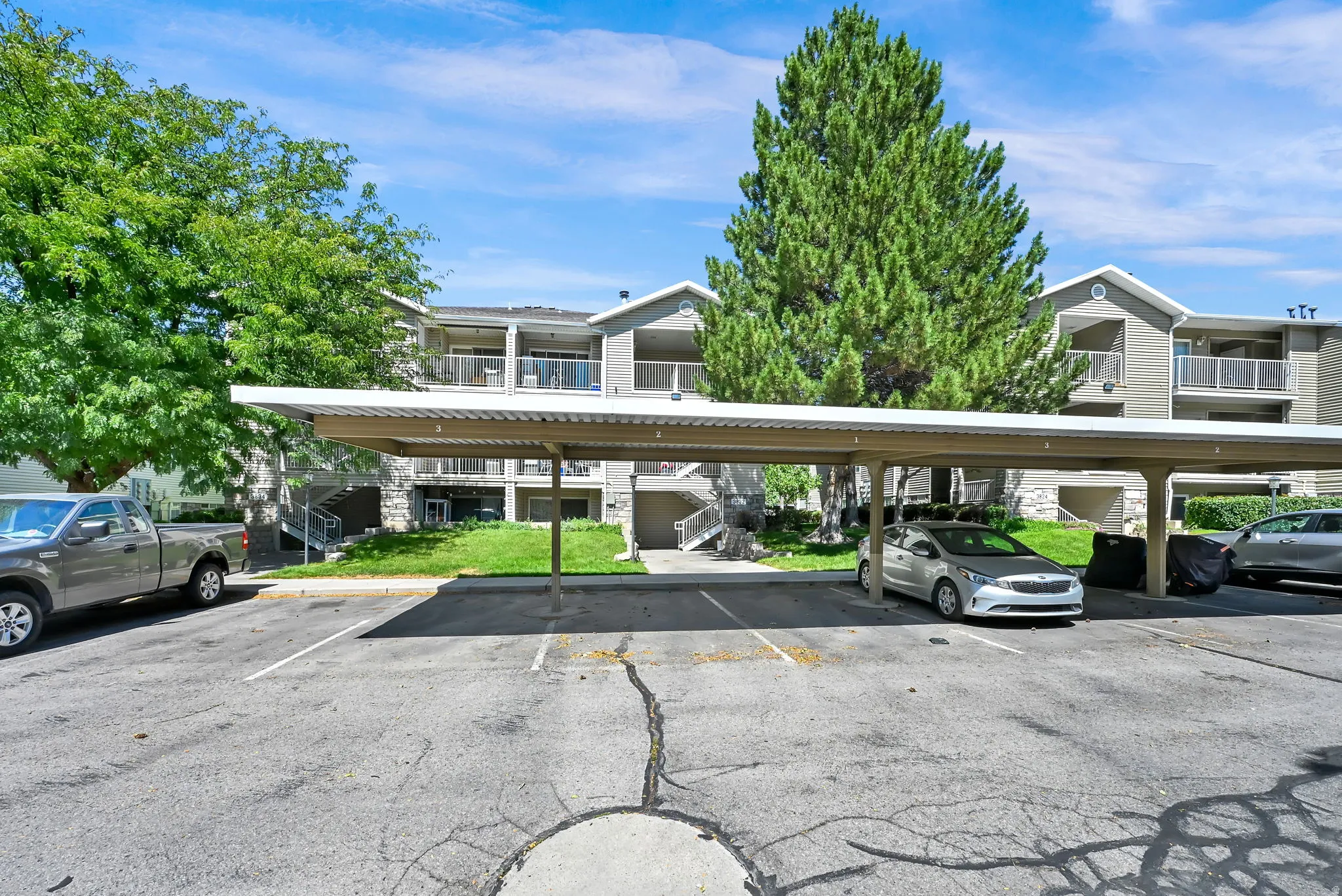 Covered parking structure with multiple spaces, situated in front of a multi-story building featuring balconies and mature landscaping