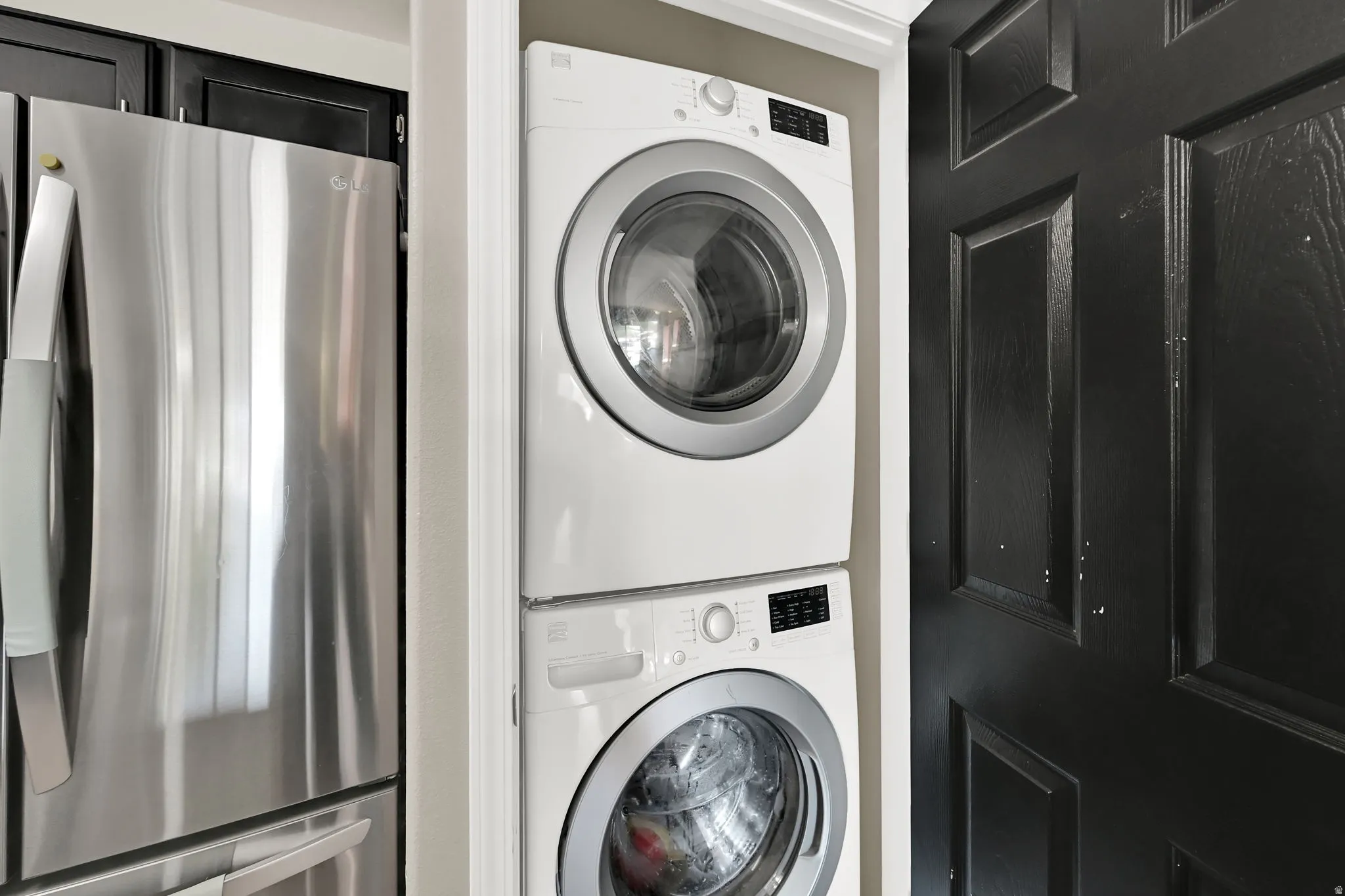 Stainless steel refrigerator, black cabinetry, white stacked washer and dryer, and a paneled black door