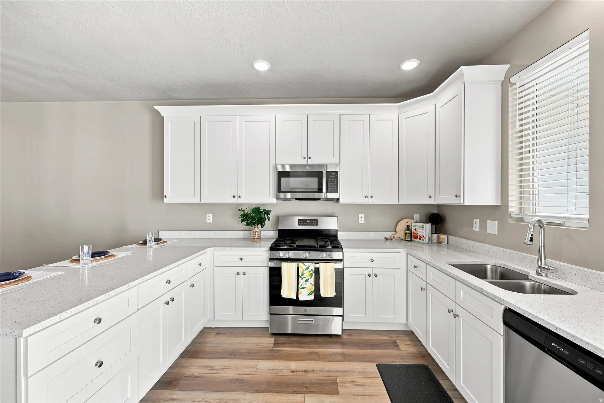 White shaker cabinetry, light-toned countertops, wood-finish flooring, stainless steel appliances, and a double basin sink