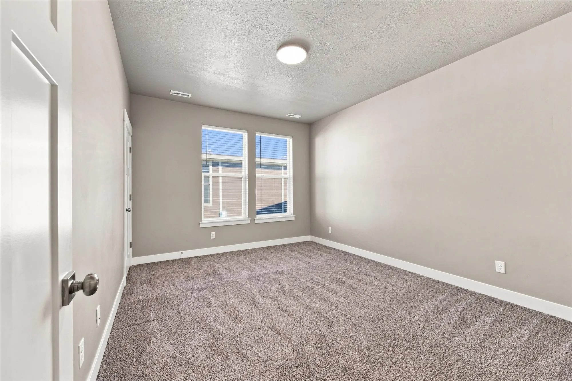 Large bedroom with new paint, textured carpeting, double windows with blinds, prominent ceiling light fixture, and white trim.