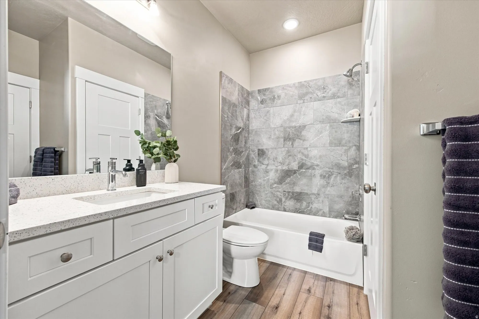 White vanity with a speckled quartz countertop and integrated sink, chrome faucet, wood-finish flooring, and a tiled shower-tub combination.