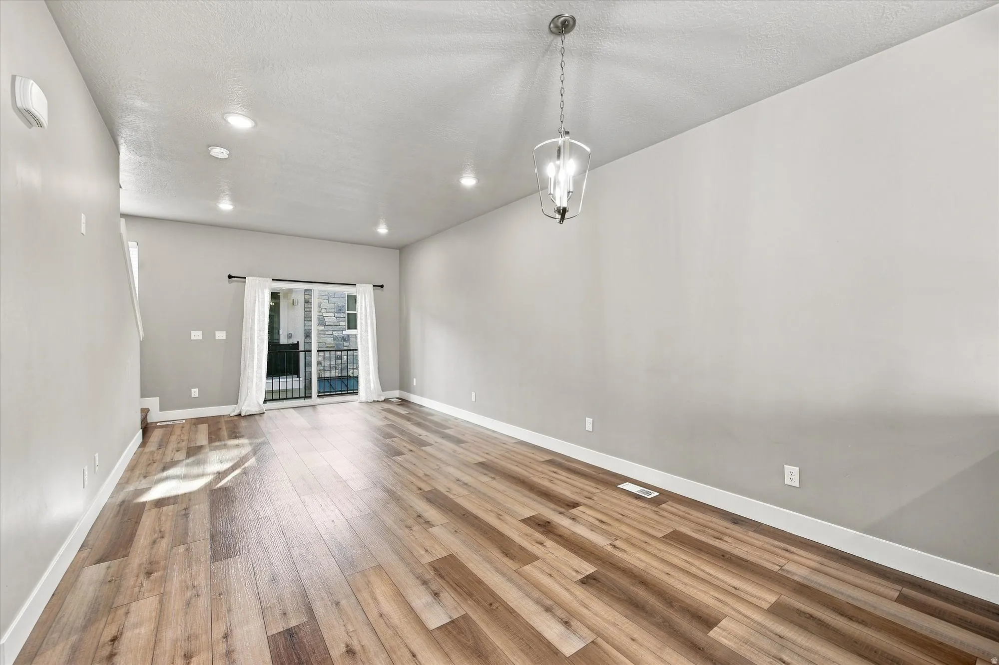 Spacious room featuring wood-finish flooring, recessed lighting, a modern chandelier, and a sliding glass door leading to a balcony.
