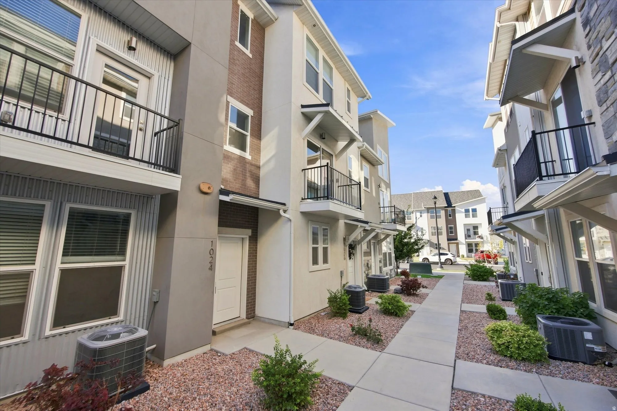 Walk way to beautiful townhomes. Modern multi-level exterior with brick and panel siding, featuring a private balcony with black railings, and a concrete pathway.
