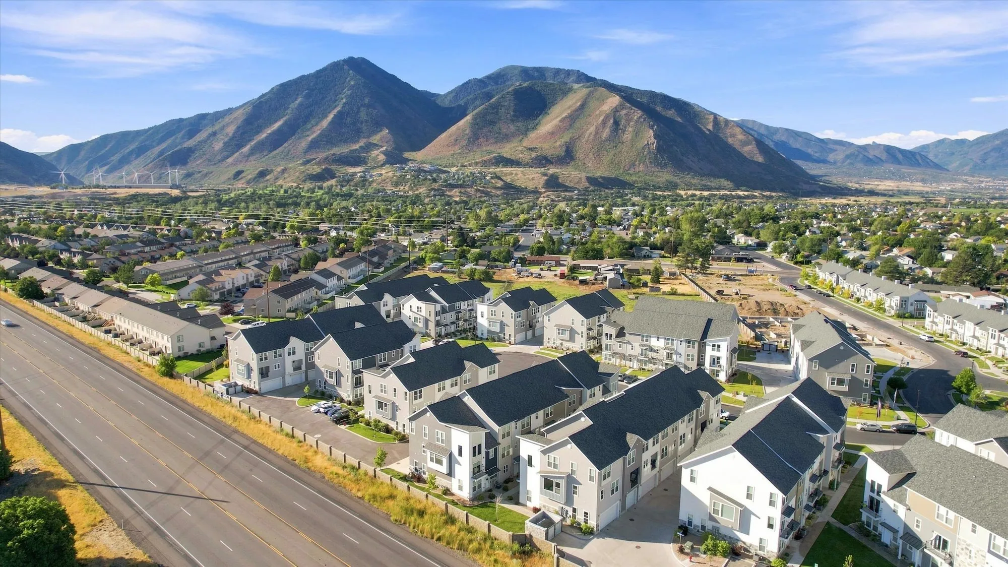 Residential development featuring multi-story townhomes, dark shingle roofs, light-toned siding, and paved roadways, set against a mountainous backdrop