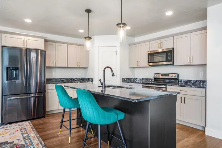 Kitchen featuring an island with a built-in sink, stainless steel appliances, light-toned cabinetry, dark countertops, and wood-finish flooring