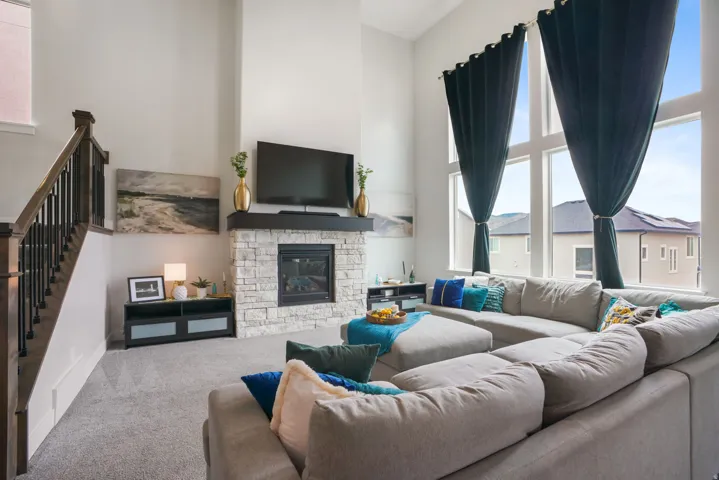Two-story ceiling, stone-clad fireplace with wood mantle, large multi-pane windows, and a staircase with wood handrails and black balusters