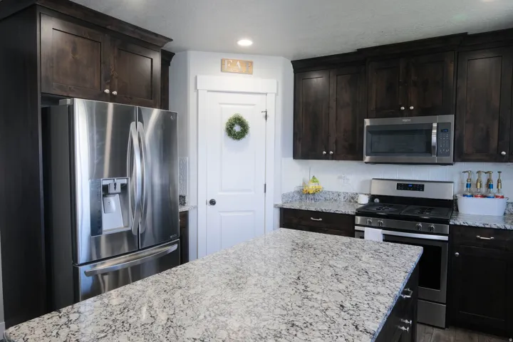 Dark wood-finish cabinetry, stainless steel appliances, light-toned stone countertops, and a white tile backsplash