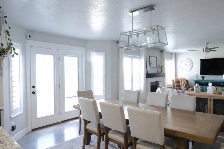 Wood-finish flooring, white double French doors, two vertical windows with plantation shutters, and a dining area with an overhead linear chandelier