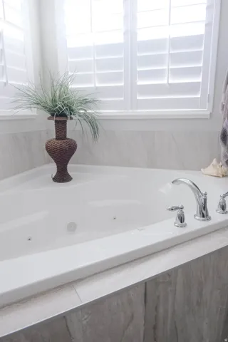 White jetted soaking tub featuring chrome fixtures, white plantation shutters, and light-toned tile surround