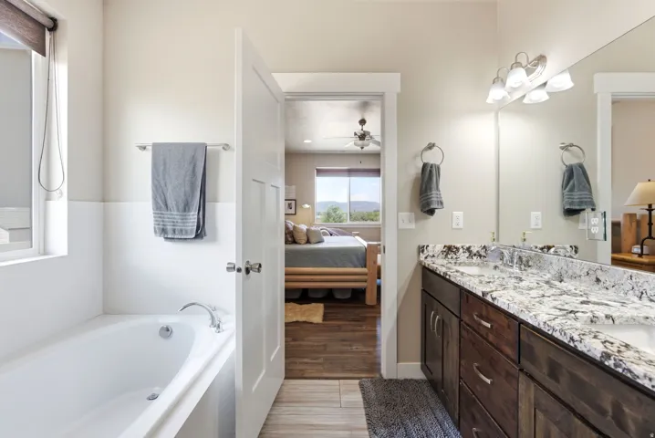 Spa-inspired primary bathroom featuring a soaking tub with tile surround, dual-sink vanity with speckled stone countertops, wood cabinetry, and abundant natural light