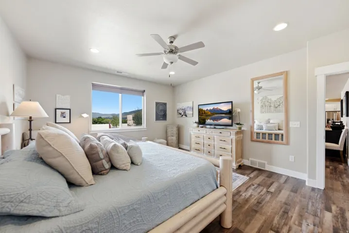 Spacious primary bedroom with large windows framing natural light, ceiling fan with integrated lighting, wood-finish flooring throughout, built-in dresser cabinetry with ample storage, and an ensuite bathroom accessible via the adjacent doorway