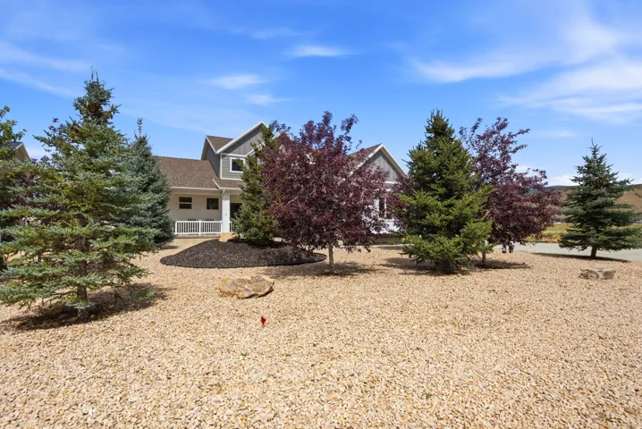 Gravel landscaping featuring diverse mature trees, with a two-story residence visible in the background, showcasing a covered porch and gabled roofline