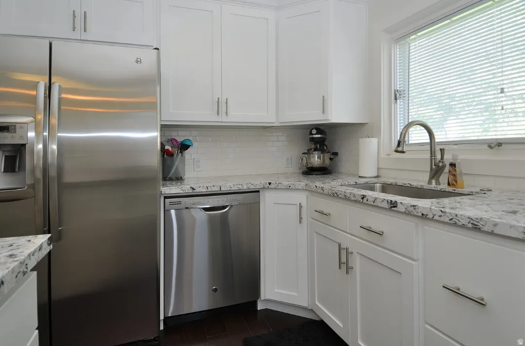White shaker-style cabinetry with brushed nickel hardware, stainless steel appliances, speckled countertops, white subway tile backsplash, and dark wood-finish flooring