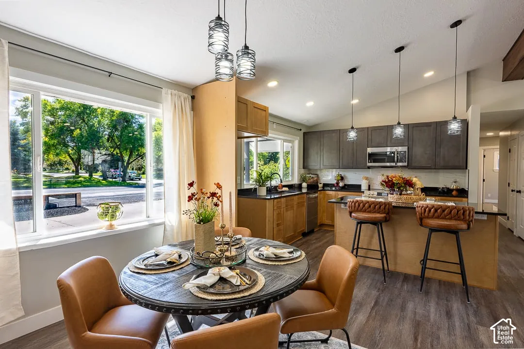Open-concept living space featuring wood-finish flooring, a kitchen island with pendant lighting, stainless steel appliances, and a vaulted ceiling
