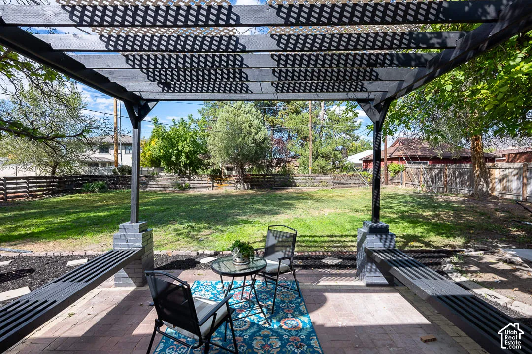 Pergola-covered patio with paver flooring, built-in benches, and direct access to a tiered, fenced backyard with mature trees