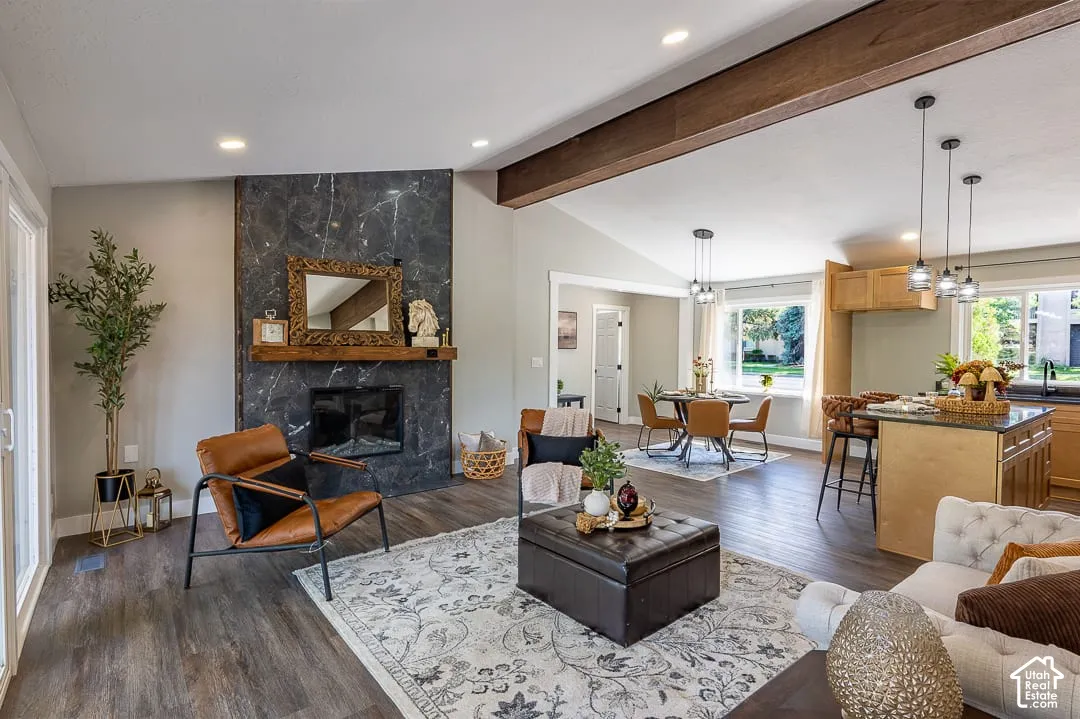 Vaulted ceiling with exposed wood beam, a black-veined stone fireplace with wood mantel, wood-finish flooring throughout, recessed lighting, and a kitchen island with pendant lighting