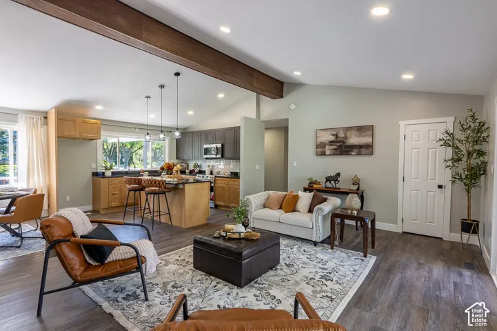 Open-concept living area featuring a vaulted ceiling with a wood beam, recessed lighting, wood-finish flooring, and a kitchen with an island and pendant lighting