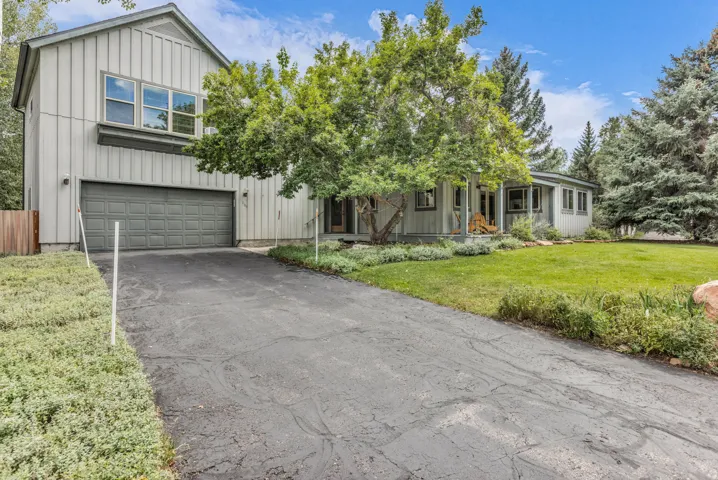 Board and batten siding, attached two-car garage, multiple windows, front yard with mature trees, and a paved driveway