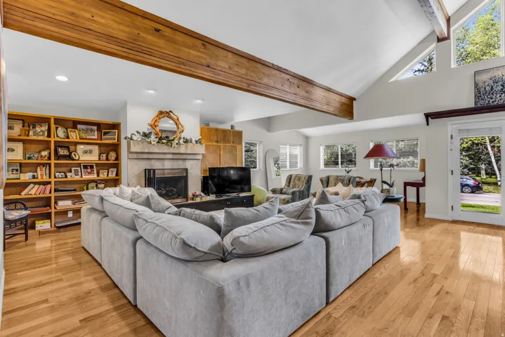 Vaulted ceiling with exposed wood beams, wood-finish flooring, stone-clad fireplace, built-in shelving, and multiple windows
